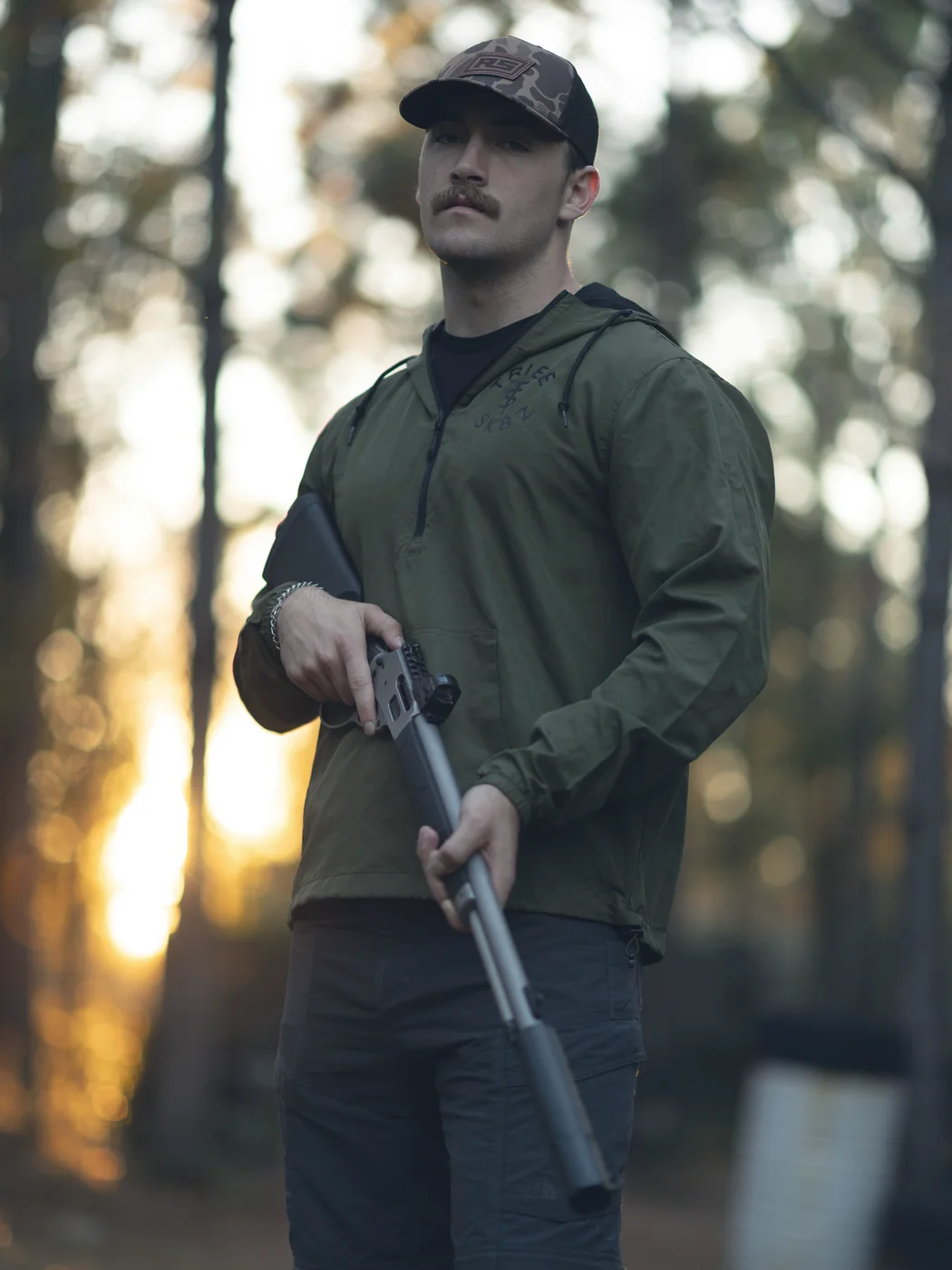Hunter in an RS cap holding a suppressed lever gun in the pines at sunset