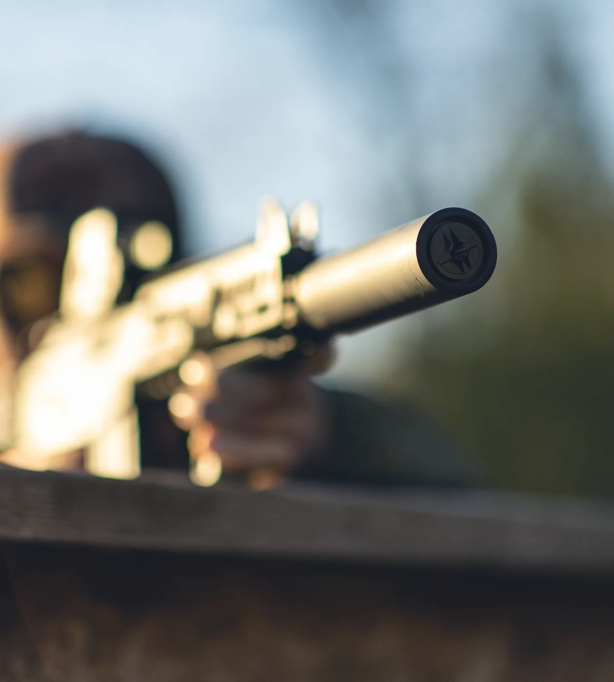 Shooter aiming a suppressed rifle over a weathered rail, endcap sharp to camera at golden hour